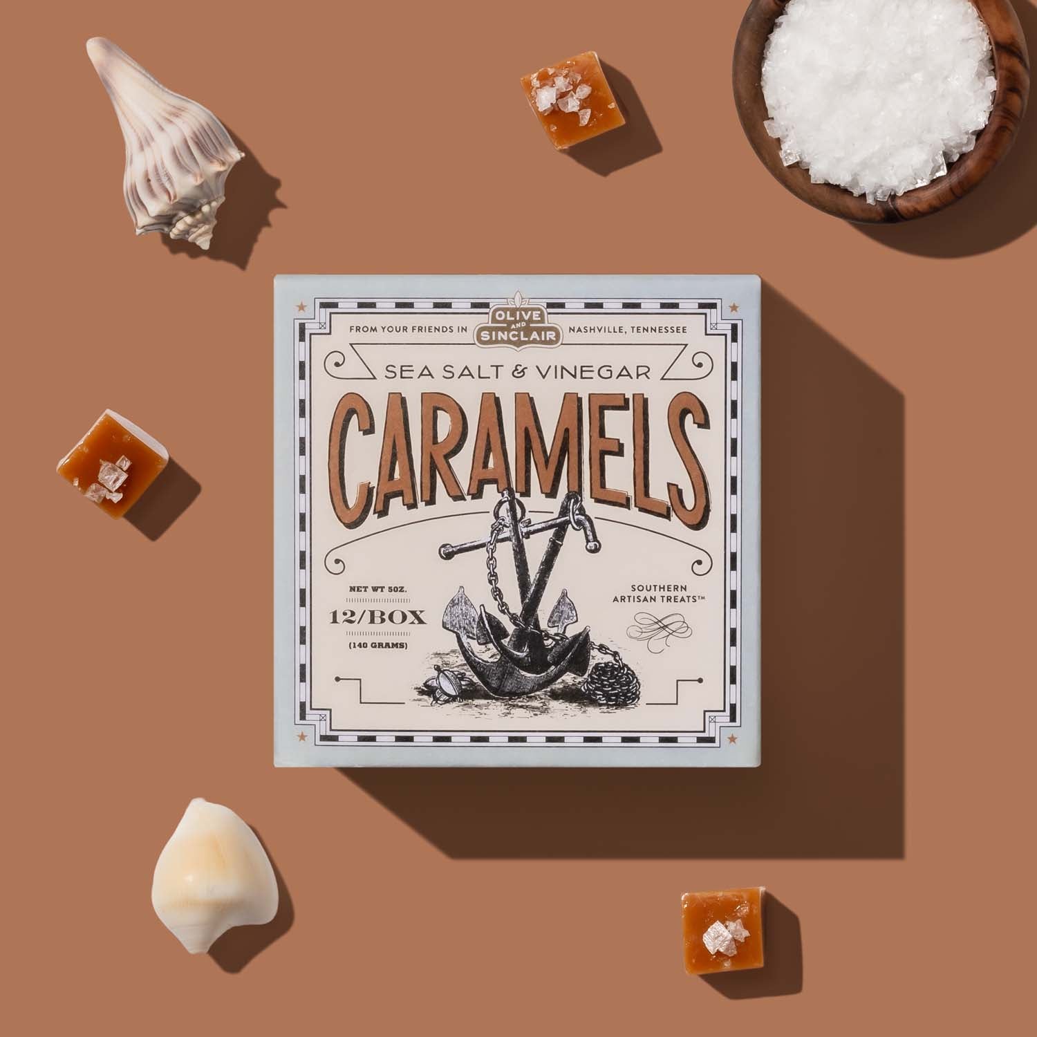 Box of sea salt and vinegar caramels on a brown background with caramels and a bowl of salt.
